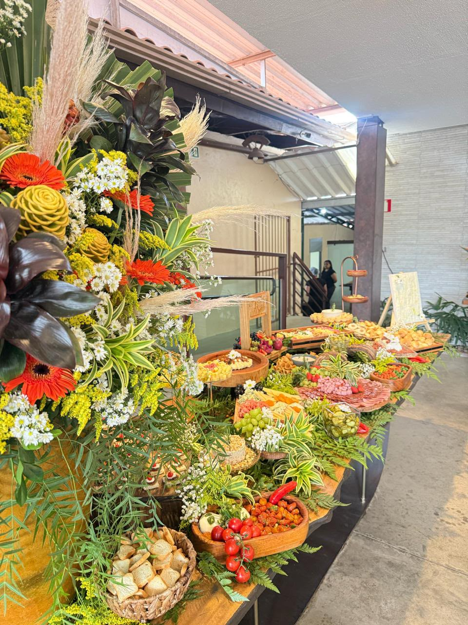 Mesa de buffet farta com queijos, frios, pães e grande arranjo floral